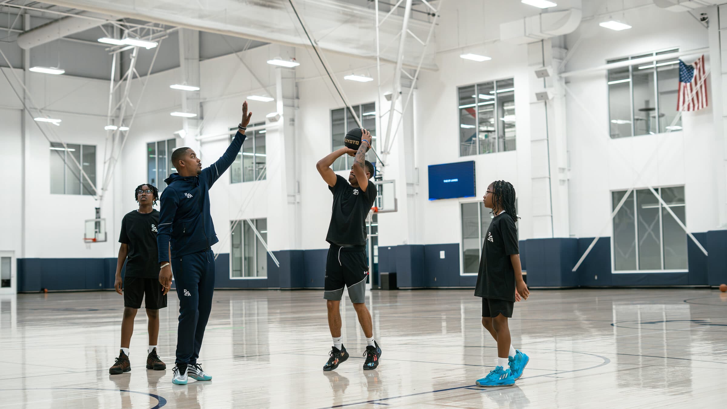 A Petty Athletics coach raises one arm beside a young player shooting a jump shot while two other young players watch in a bright indoor gym